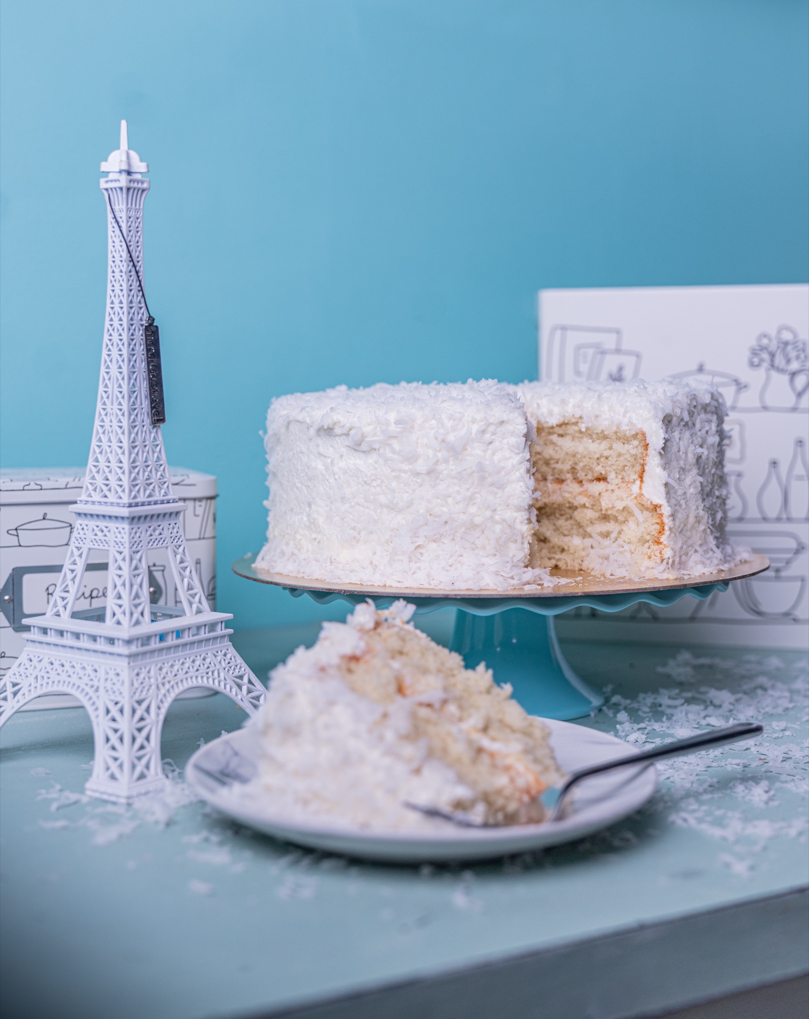 A fully iced coconut cake with a slice cut away, styled beside a small Eiffel Tower model